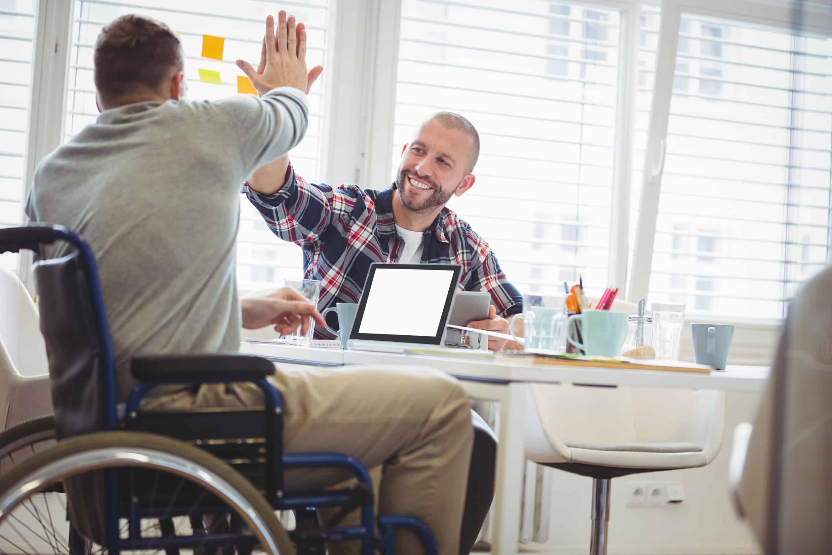 Ein Mann sitzt mit dem Rücken zu uns in einem Rollstuhl. Vor ihm steht ein Computer. Ihm gegenüber sitzt ein junder Mann. Die Beiden klatschen über dem Computer ihre rechten Hände zusammen und freuen sich.