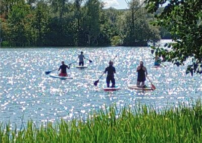 Im Sonnenschein sind vier Menschen auf einem Stand-Up-Paddling-Board auf einem See zu sehen. Sie knien auf dem Board und bewegen sich paddelnd fort.