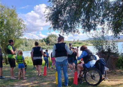 Eine Gruppe von Menschen steht an einem See. Alle tragen Schwimmweszem- Ein Mann sitzt im Rollstuh. Ihm wird gezeigt, wie er ein Paddel zu benutzen hat. Alle warten darauf über einen Steg zu Stand-Up-Paddling-Boards zu gelangen