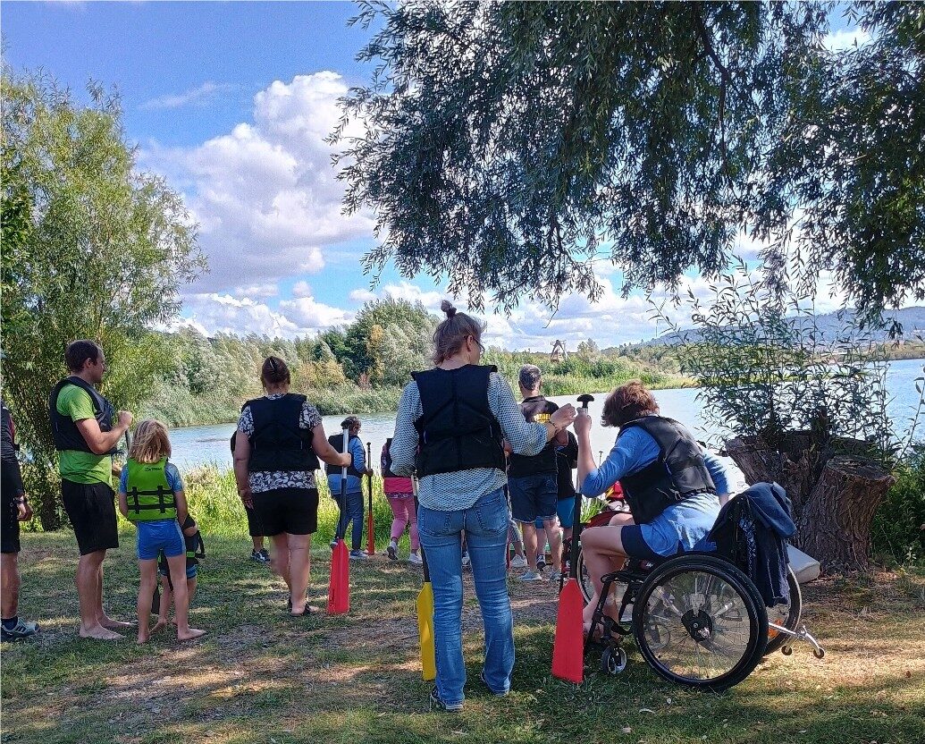 Paddeln für Menschen mit und ohne Behinderung Eine Gruppe von Menschen steht an einem See. Alle tragen Schwimmweszem- Ein Mann sitzt im Rollstuh. Ihm wird gezeigt, wie er ein Paddel zu benutzen hat. Alle warten darauf über einen Steg zu Stand-Up-Paddling-Boards zu gelangen